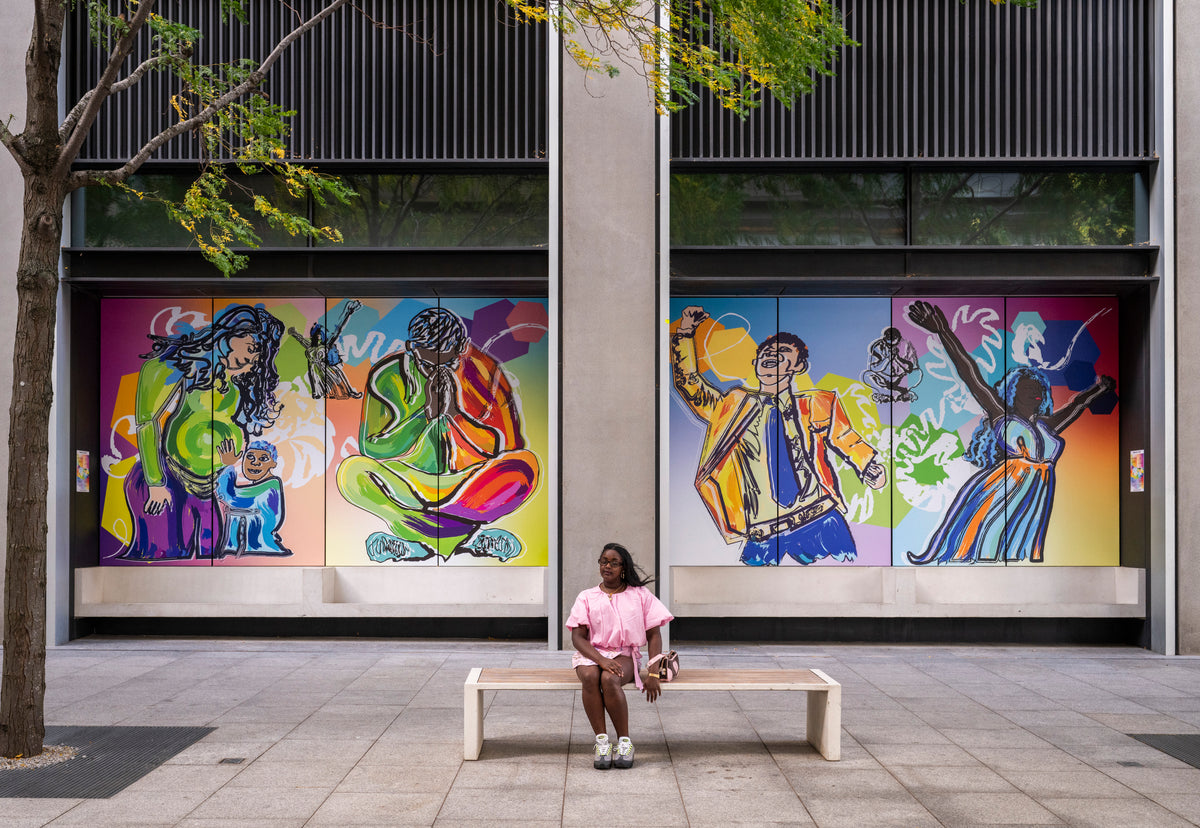 A woman sits on a white bench in front of a modern building facade featuring two large colorful mural windows with stylized figures. The scene is outdoors on a paved plaza with a tree on the left.